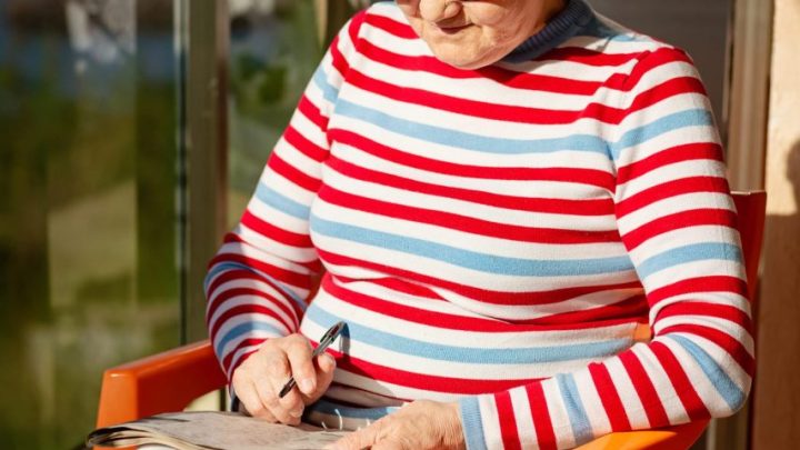 Personne âgée faisant des mots mêlés à gros caractères sur une feuille imprimée, assise à une table avec un stylo.